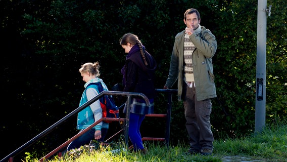 Ein Mann hält seinen Zeigefinger an den Mund und deutet Ruhe an. Vor ihm gehen Jessi und Nina eine Treppe hinunter. © NDR Foto: Claudia Timmann