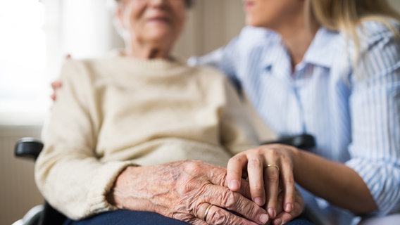 Eine Pflegerin legt den Arm um eine alte Frau im Rollstuhl und hält deren Hand. © Colourbox Foto: 232319