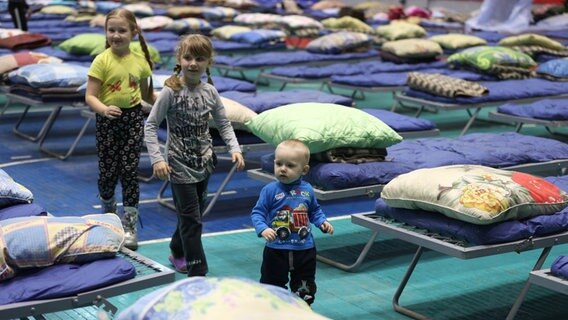 Drei Kinder gehen durch eine Flüchtlingsunterkunft mit in Reihen aufgestellten Feldbetten © picture alliance/dpa/TASS | Erik Romanenko Foto: picture alliance/dpa/TASS | Erik Romanenko