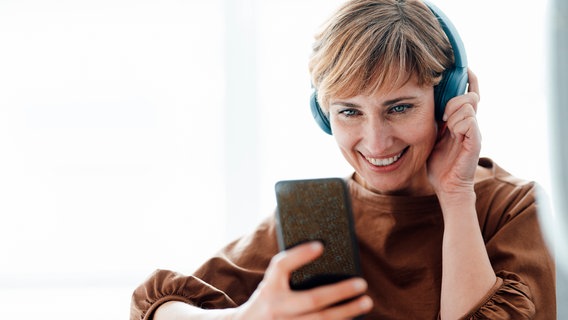 Ein Frau, die Kopfhörer trägt, schaut lächelnd auf ihr Smartphone. © imago images Foto: Westend61