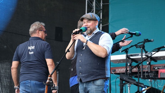 Stefan Kuna von NDR 1 Radio MV steht mit Schiebermütze und Mikrofon auf der Bühne © NDR Foto: NDR