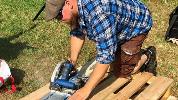 Ein Mann mit Sonnenbrille sägt eine Palette mit einer elektrischen Säge © NDR Foto: Jan Baumgart