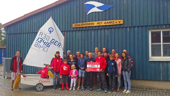 Mitglieder des Seglerverein Alt Gaarz e.V. stehen vor einem Vereinshaus und präsentieren einen 1.000-Euro-Scheck. © NDR Foto: Steffen Baxalary