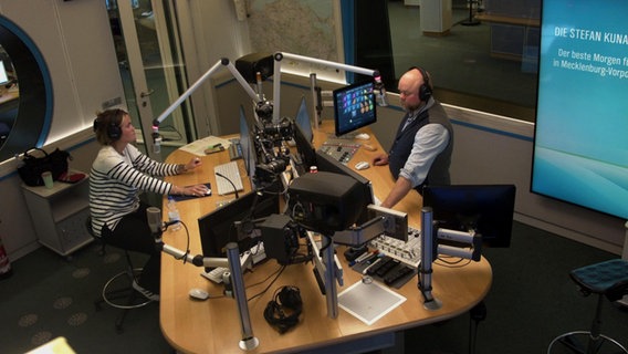 Webcamansicht eines NDR 1 Radio Mv Studios mit Mischpulten, Mikrofonen und Monitoren. Stefan Kuna und Theresa Hebert sitzen im Studio © ndr Foto: ndr