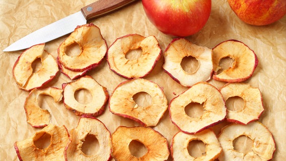 Getrocknete Apfelscheiben, zwei Äpfel und eine Messer liegen auf einem Stück Backpapier. © Colourbox 