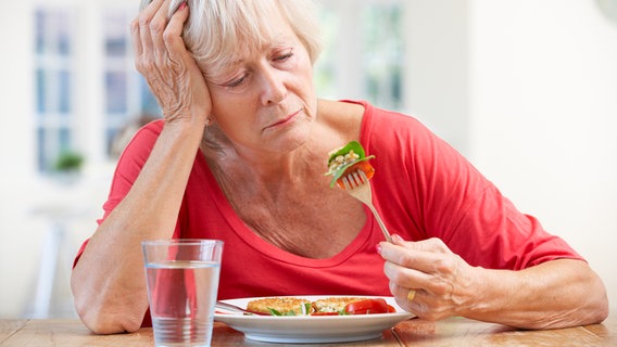 Eine ältere Frau sitzt vor einem Teller mit Essen und führt eine Gabel zum Mund. © colourbox 