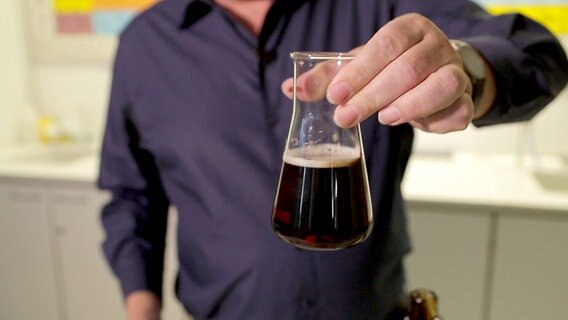 Hand hält eine Kolbenflasche in der Hand, die mit dunklem Bier gefüllt ist. © NDR 