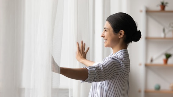 Eine Frau steht am Fenster und schiebt weiße Gardinen beiseite. © Colourbox Foto: Aleksandr