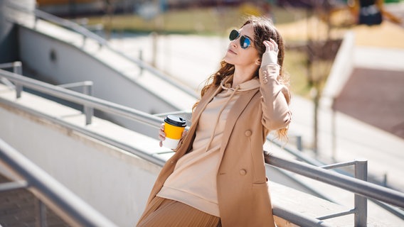 Eine Frau steht mit einem Kaffeebecher an einem Geländer in der Sonne. © Colourbox Foto: Mykola