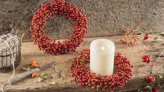 Eine brennende Kerze steht in der Mitte eines Kranzes aus Hagebutten © imago images / imagebroker 