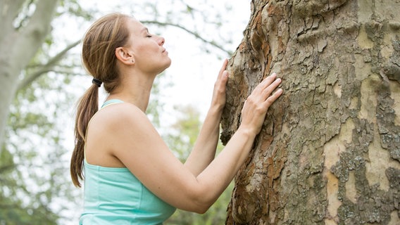 Eine Frau steht mit geschlossenen Augen an einem Baum, den sie mit ihren Händen berührt. © picture alliance / dpa-tmn | Christin Klose 