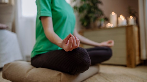 Eine Frau beim Yoga. Sie sitzt in der Lotus-Position auf einem Kissen. © picture alliance Foto: picture alliance / Westend61 | HalfPoint