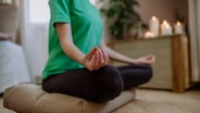 Eine Frau beim Yoga. Sie sitzt in der Lotus-Position auf einem Kissen. © picture alliance Foto: picture alliance / Westend61 | HalfPoint