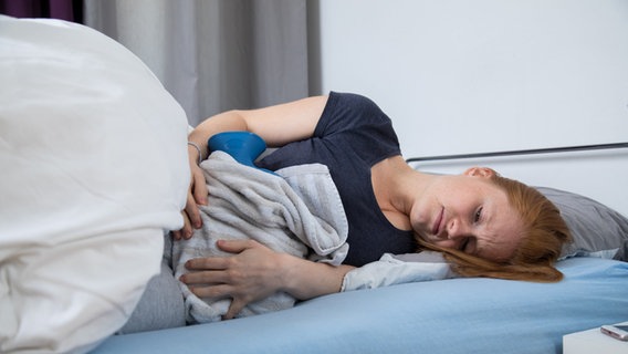 Eine junge Frau liegt in einem Bett und leidet unter Bauchschmerzen.  © picture alliance / dpa-tmn |  Christine Klose 
