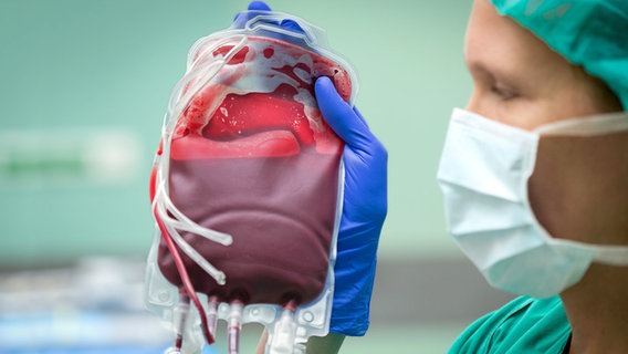 Eine Frau in OP-Kleidung hält eine Blutkonserve (durchsichtiger Beutel mit Blut) hoch. © Friso Gentsch/dpa Foto: Friso Gentsch