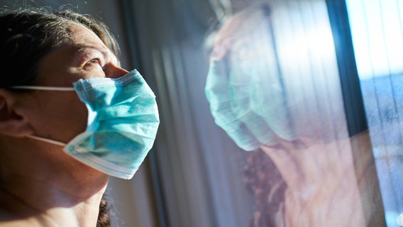 Eine Frau mit Mundschutz steht an einem Fenster und blickt ins Licht. © imago images Foto: Action Pictures/imago