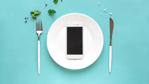 Ein Handy liegt auf einem Teller mit Besteck daneben und ein paar Kräutern auf dem Tisch. © Colourbox Foto: #228088