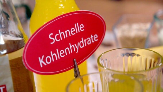 Vor einer Saftflasche steht ein Schild mit der Aufschrift "Schnelle Kohlenhydrate". © NDR 