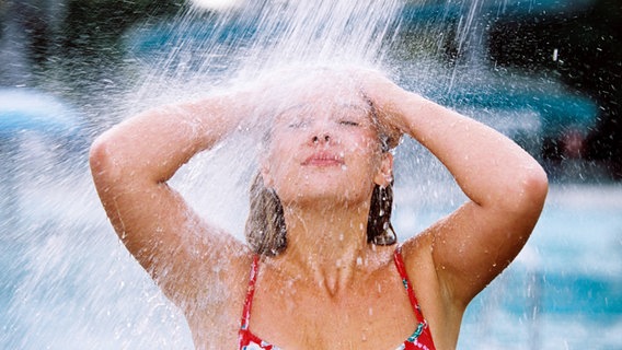 Eine Frau im Bikini erfrischt sich unter einer Dusche im Freien. © imago/imagebroker/obermeier 