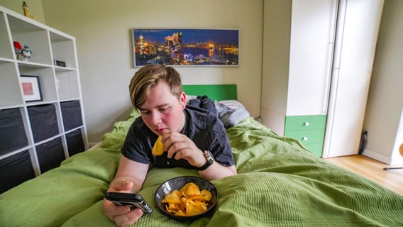 Ein Jugendlicher liegt im Bett, schaut aufs Handy und isst Chips. © NDR Foto: Moritz Schwarz / Oliver Zydek