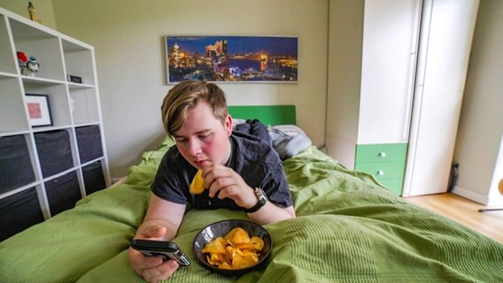Ein Jugendlicher liegt im Bett, schaut aufs Handy und isst Chips. Ein Jugendlicher liegt im Bett, schaut aufs Handy und isst Chips. © NDR Foto: Moritz Schwarz / Oliver Zydek
