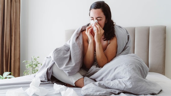 Eine kranke Frau sitzt auf ihrem Bett und putzt sich die Nase. © Imago Images / Westend61 Foto: Ok Shu