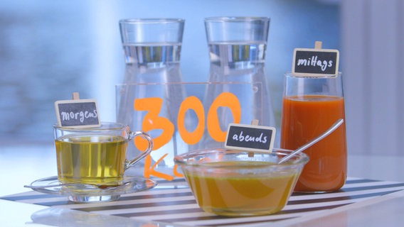 Wasser, Tee, Gemüsesaft und Fastenbrühe stehen auf einem Tisch, daneben das Schild "300 Kcal". © NDR 