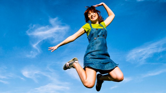 Lächelnde Frau mit roten Haaren beim Sprung in der Luft vor blauem Himmel. Sie trägt ein kurzes Jeanskleid mit T-Shirt darunter. © Colourbox.de/Erik Reis 