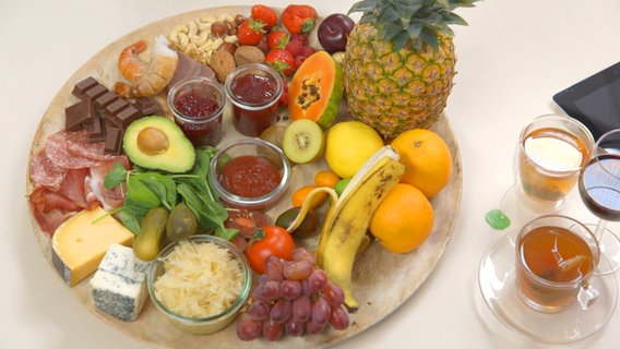 Ein großer Teller mit verschiedenen Nüssen, Erdbeeren, Bananen, Tomaten, Essiggurken, Ketchup, Blauschimmelkäse, Papaya, Ananas, Sauerkraut, Zitrusfrüchten, Kiwi, Schokolade, Marmelade, Krustentieren und Pflaumen, dazu grüner Tee und Rotwein.  ©NDR 