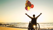 Eine Frau sitzt im Sonnenuntergang am Meer in einem Rollstuhl, an dem mehrere Luftballons befestigt sind. © colourbox Foto: Ikostudio