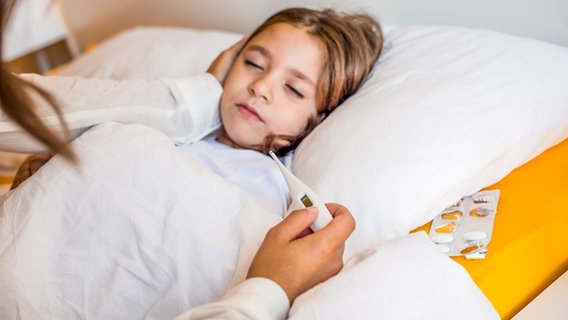 Eine Frau sitzt am Bett eines kranken Kindes und hält ein Fieberthermometer in der Hand © Colourbox 