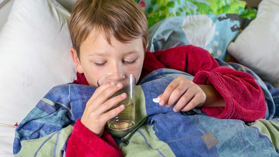 Ein kranker Junge liegt im Bett, trinkt aus einem Glas und hält eine Tablette in einer Hand © Colourbox 