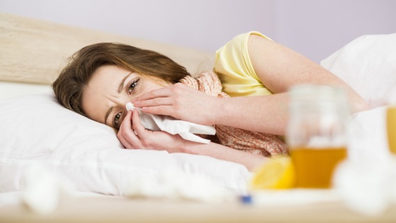 Eine kranke Frau liegt im Bett und putzt sich die Nase. © Colourbox 
