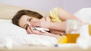 Eine kranke Frau liegt im Bett und putzt sich die Nase. © Colourbox 