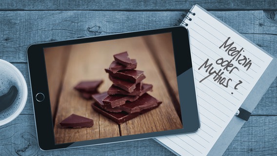 Auf einem Tisch liegt ein Tablet mit einem Bild von dunkler Schokolade auf einem Holzbrett. Auf einem Notizblock sind die Worte "Medizin oder Mythos" zu lesen (Montage) © Colourbox/Photocase Foto: Blackzheep/Gortincoiel