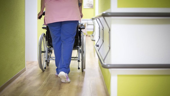 Eine Altenpflegerin schiebt eine Bewohnerin in einem Altenpflegeheim in einem Rollstuhl den Gang entlang © imago Foto: photothek