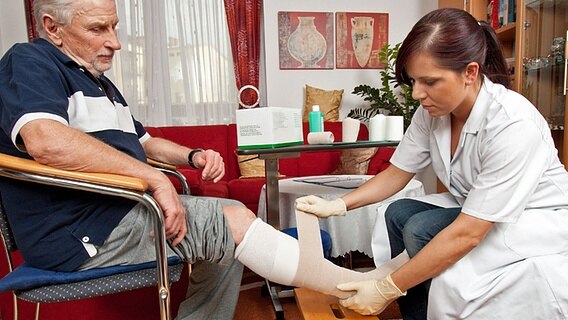 Eine Pflegerin legt einem alten Mann einen Verband am Bein an. © imago images / blickwinkel/McPhoto 