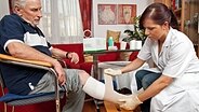 Eine Pflegerin legt einem alten Mann einen Verband am Bein an. © imago images / blickwinkel/McPhoto 