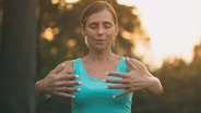 Eine Frau steht in der Dämmerung in der Natur und führt eine Qi Gong Übung aus. Die Hände sind vor ihrer Brust angehoben. © IMAGO / Depositphotos 