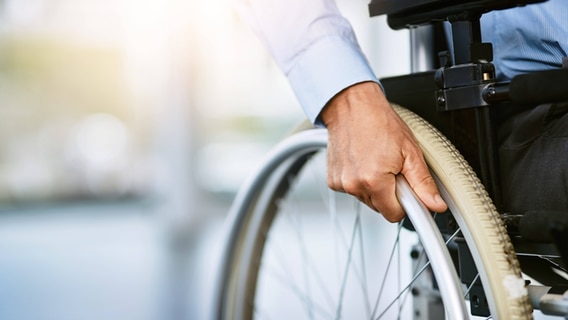 Die Hand eines Mannes am Rad eines Rollstuhls. © IMAGO / Zoonar II 
