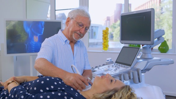 Dr. Jörn Klasen sitzt neben der Patientin Charline W., die auf einer Liege liegt, und untersucht ihre Schilddrüse. © NDR 