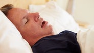 Ein schlafender Mann liegt mit offenem Mund und auf dem Rücken in einem Bett © colourbox 