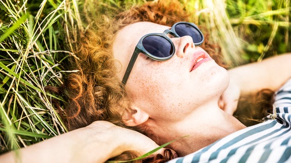 Eine junge Frau mit Sonnenbrille liegt im Gras und sonnt sich. © picture alliance / Westend61 Foto: Jo Kirchherr