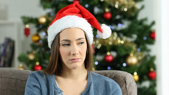 Eine Frau mit Weihnachtsmann-Mütze sitzt traurig auf einem Sofa, im Hintergrund ein geschmückter Weihnachtsbaum © colourbox 
