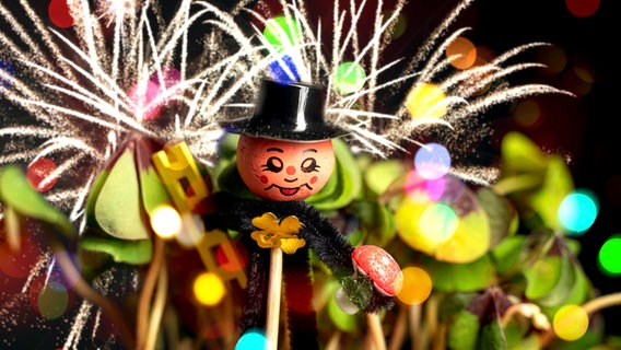 Fotomontage: Eine Schornsteinfeger-Figur steckt als Glücksbringer in einer Klee-Pflanze, oben ist ein Feuerwerk zu sehen. © picture alliance / CHROMORANGE Foto: Michael Bihlmayer