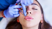 Das Gesicht einer Frau, die sich gerade mit geschlossenen Augen von einer weiteren Person mit einer Spritze Hyaluron in die Lippen spritzen lässt. © picture alliance / Shotshop | Addictive Stock 