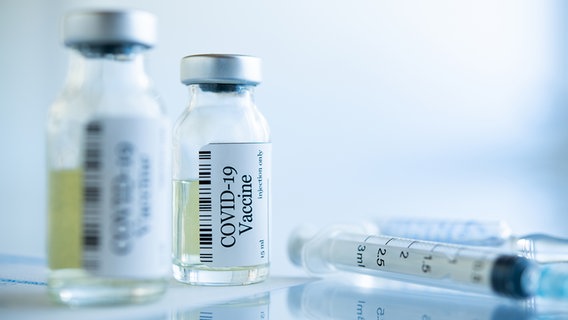 Zwei Fläschen mit Covid-19-Impfstoff und Spritzen liegen auf einem Glastisch. © PantherMedia Foto: Ridofranz