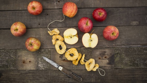 Äpfel, ein Messer und getrockente Apfelringe liegen auf auf Holztisch © imago/Westend61 