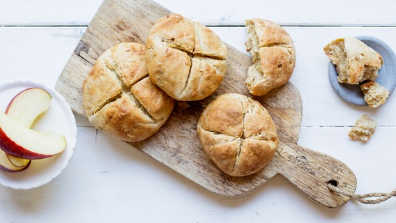 Drei Dinkel-Quark-Brötchen auf einem Holzbrett sowie daneben ein angebrochenes Dinkelbrötchen und ein angeschnittenem Apfel auf einem Teller. © NDR Foto: Claudia Timmann