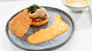 Gedämpftes Wolfsbarsch-Röllchen mit Oliven-Kräuter-Füllung, Paprikaschaum und Manchego-Segel auf einem Teller angerichtet. © NDR / Die Fernsehmacher Foto: Markus Hertrich